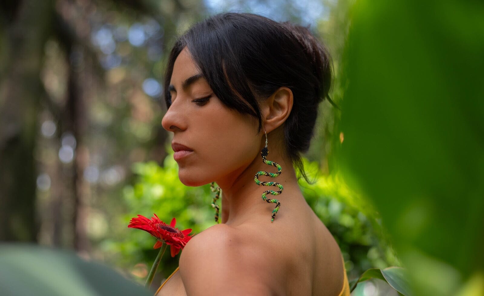 woman wearing green black gold slithering snake beaded earrings holding red flower