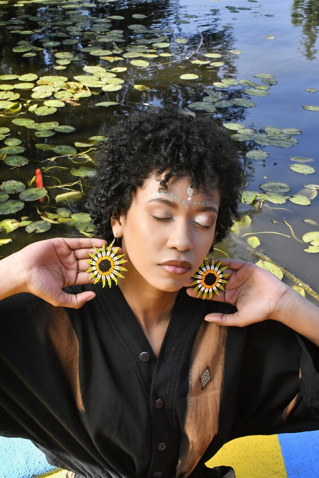 woman wearing green yellow white flower blossom pinwheel round beaded earrings by pond with lily pads
