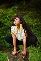 Woman wearing long yellow beaded earrings with long black hair crouching on tree trunk